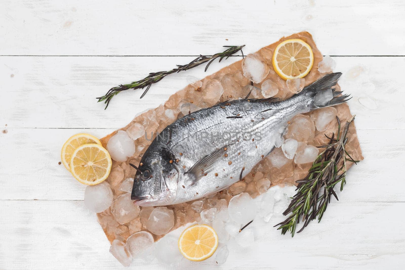 Raw dorada fish or gilt-head bream on ice with lemon slices and rosemary over white wooden background, flat lay, top view.