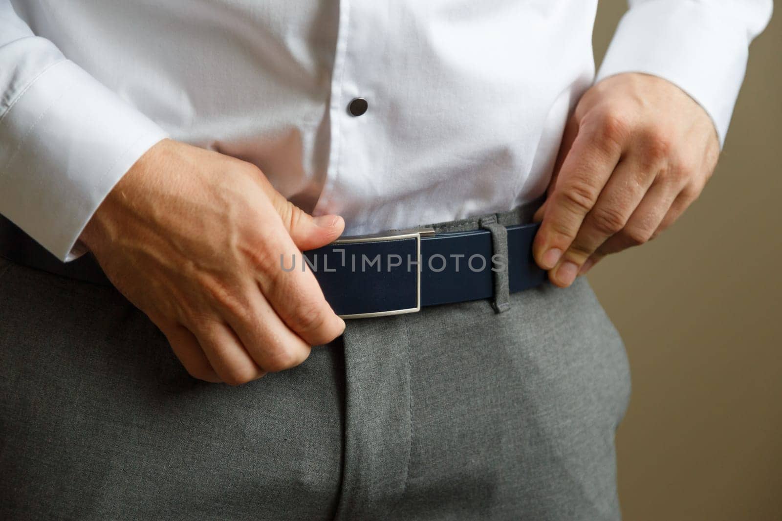 A man in blue pants and a white shirt buttoned a brown leather trouser belt. He has a watch on his hand.