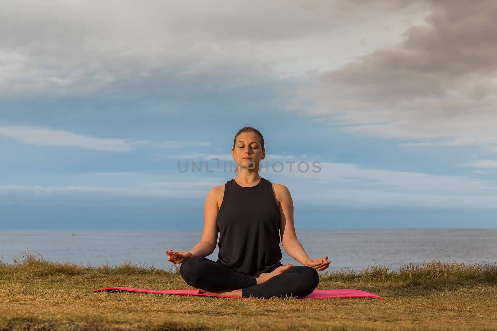 Middle aged woman in black tank top sitting on a mat doing meditation outdoors with sea and cloudy sky in background. Caucasian woman relaxing in yoga pose.