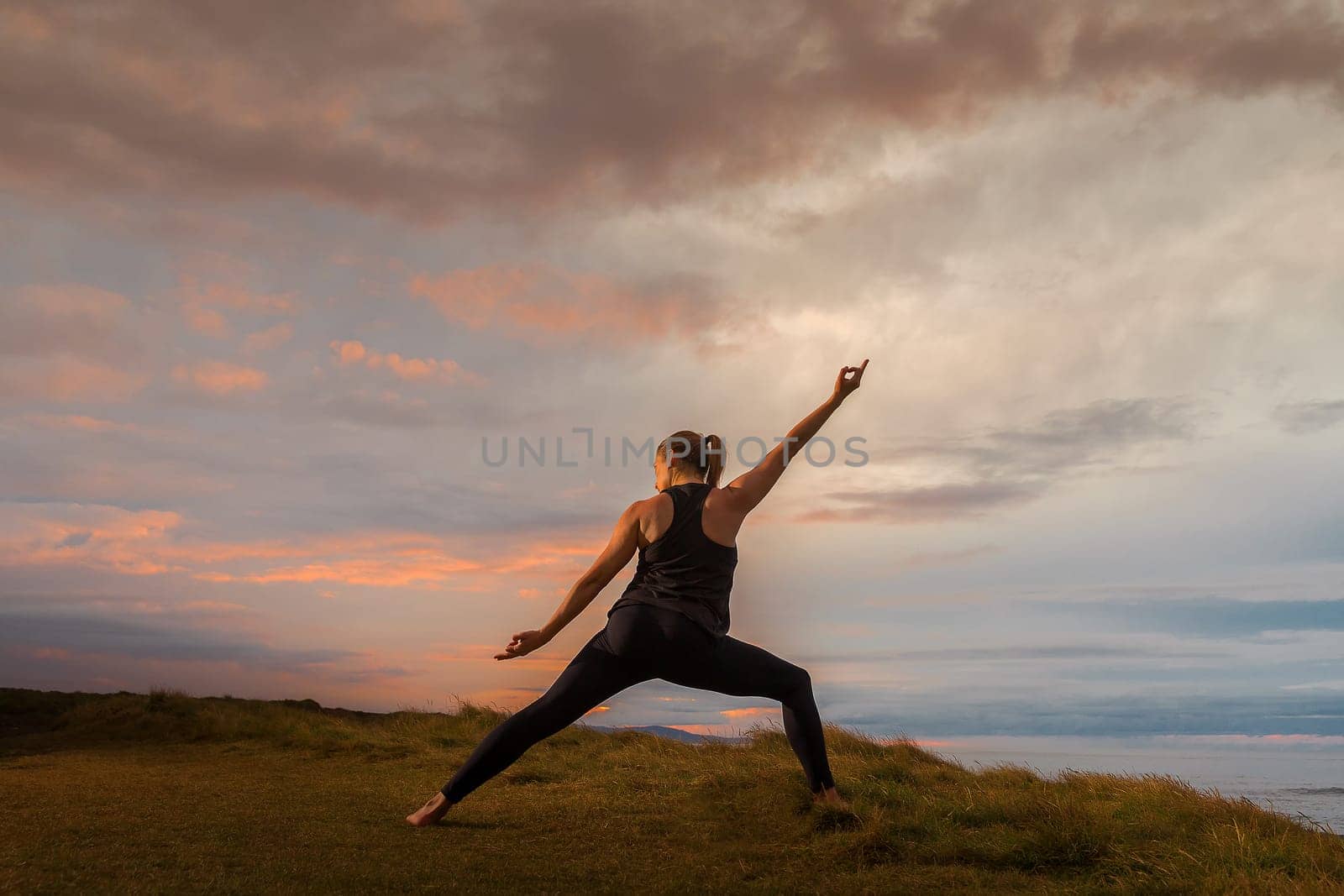 Middle-aged woman in black sportswear on her back doing meditation in front of the sea at sunset. Caucasian woman in yoga pose at dusk.