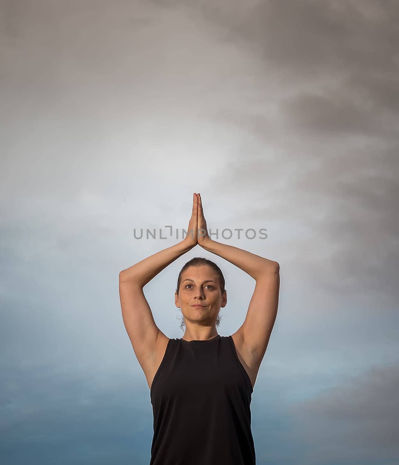 Middle-aged woman in black shirt with her arms over her head doing meditation outdoors with cloudy sky in the background. Caucasian woman relaxing in yoga pose