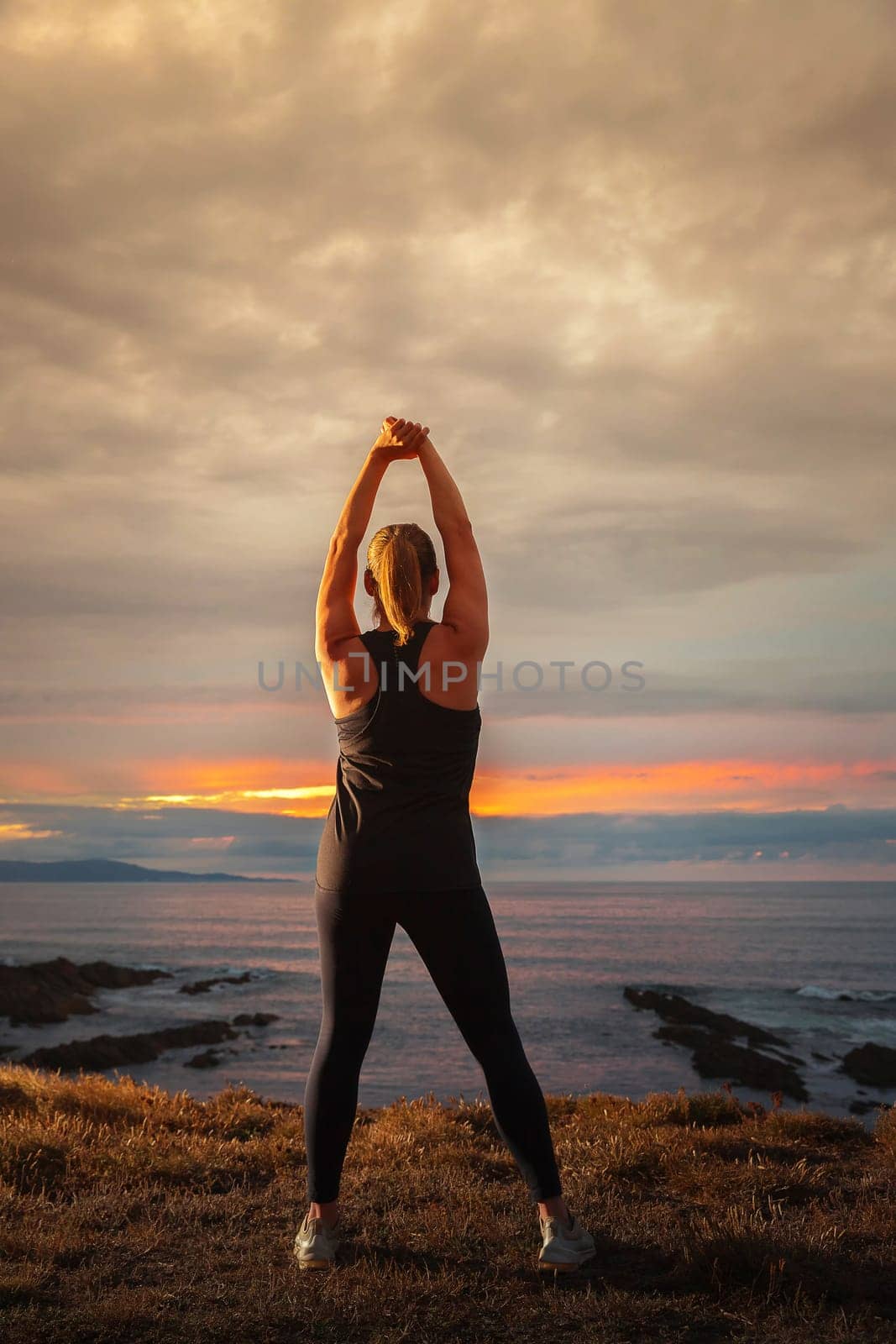 Middle-aged woman in black sportswear on her back stretching in front of the sea at sunset. Caucasian woman relaxing at dusk.