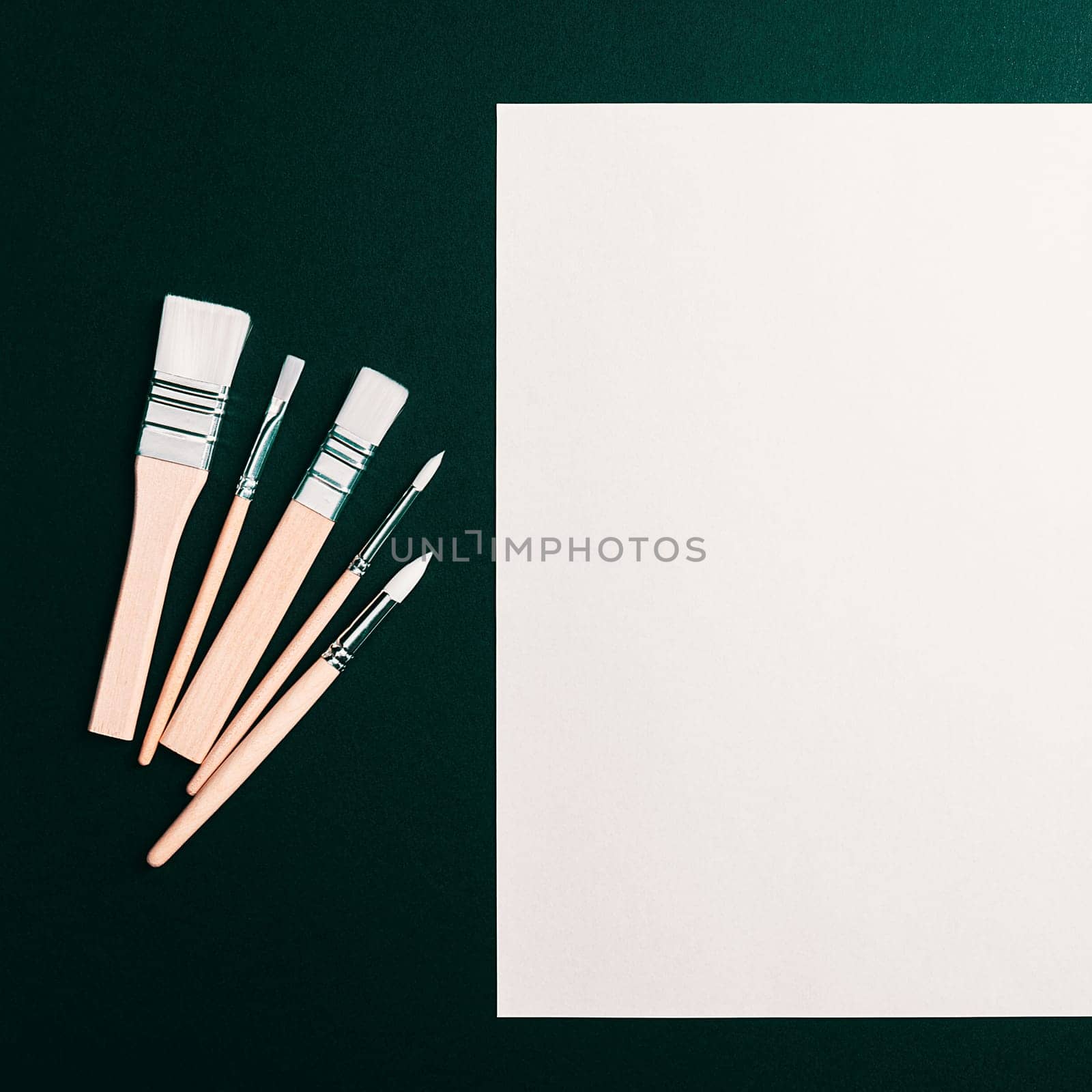 A clean white sheet and paint brushes on a dark green background with space to copy. Mock-up, mockup, layout.