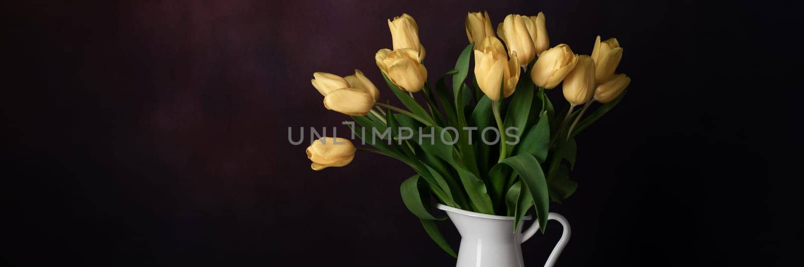 Tulips in a jug. Classic still life with a bouquet of yellow tulip flowers in a vintage white jug on a dark background and an old wooden table. Art photography.
