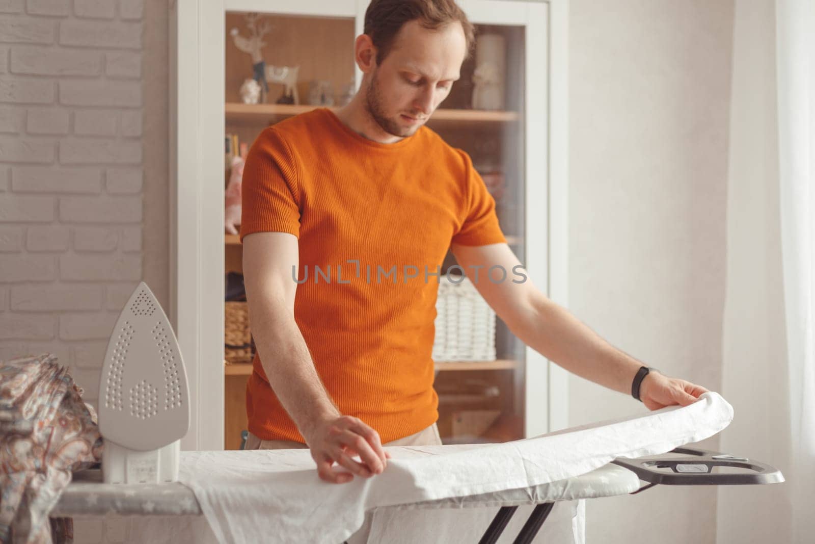 Young caucasian man ironing children's sheet on ironing board at home. Men doing home chores