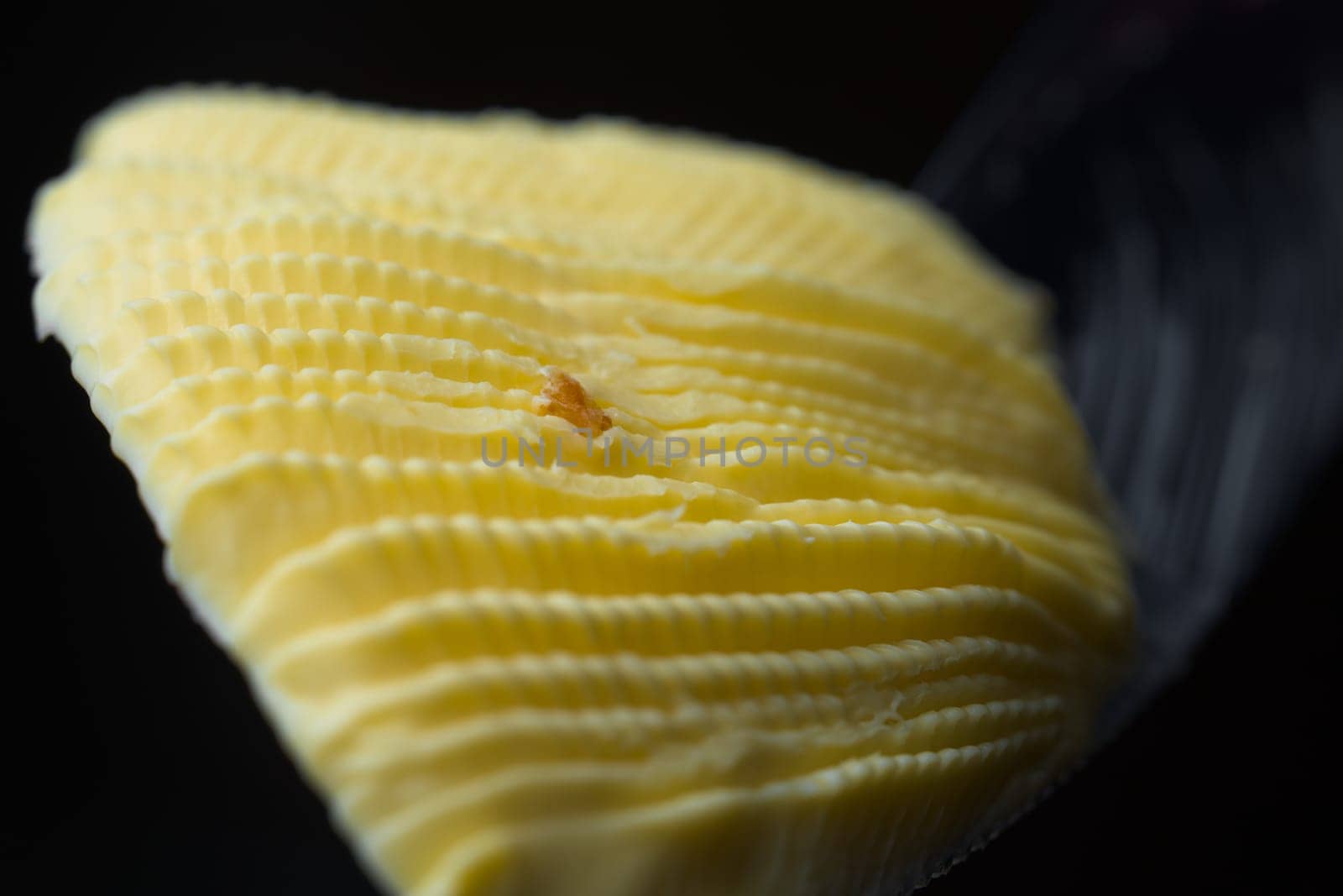 Serrated knife holds a fan shaped curl of rippled yellow margarine. Isolated on black background and a super macro perspective. Center bread crumb too