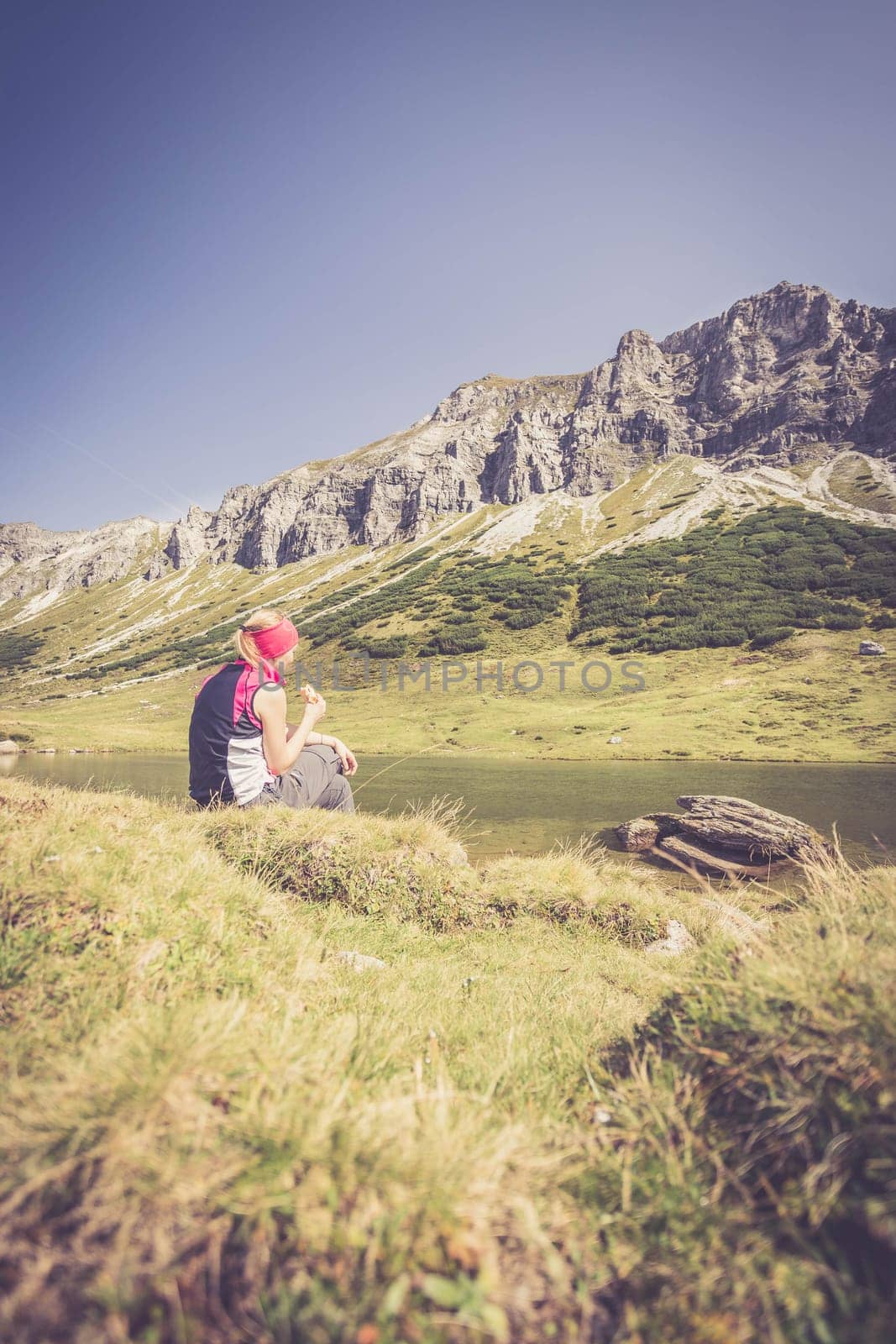 Woman in sportswear is doing a break during a hike: sitting on the ground and enjoying the view over the mountains and a lake