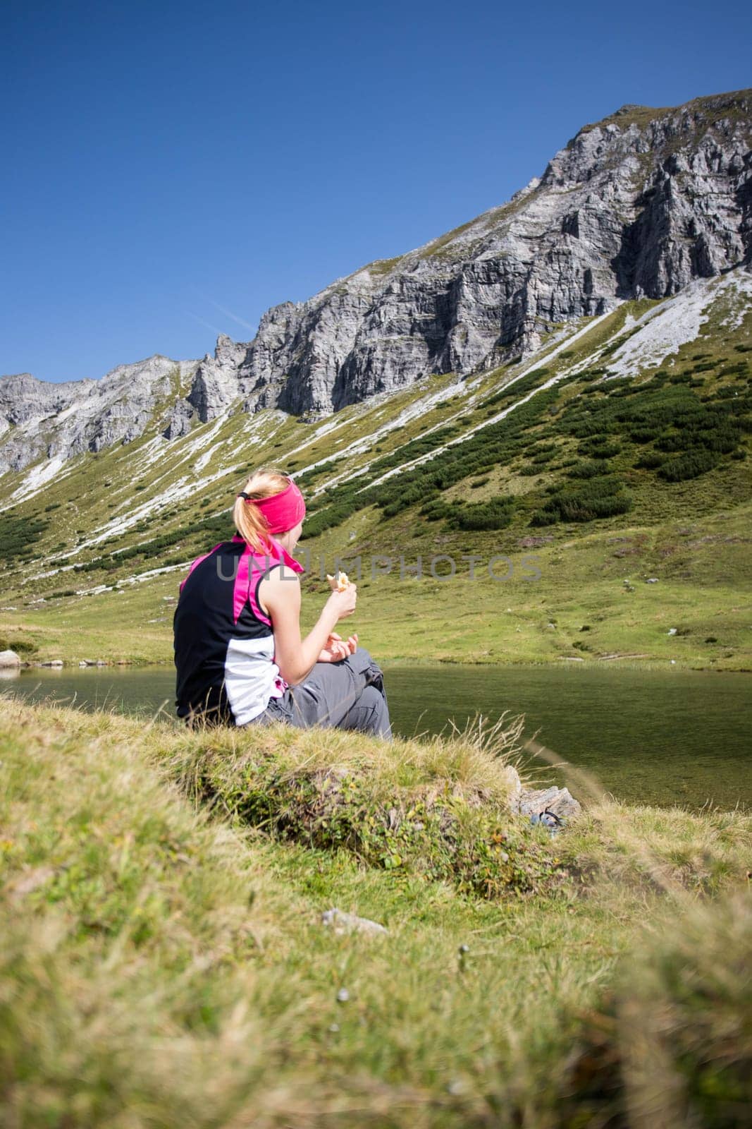 Woman in sportswear is doing a break during a hike: sitting on the ground and enjoying the view over the mountains and a lake