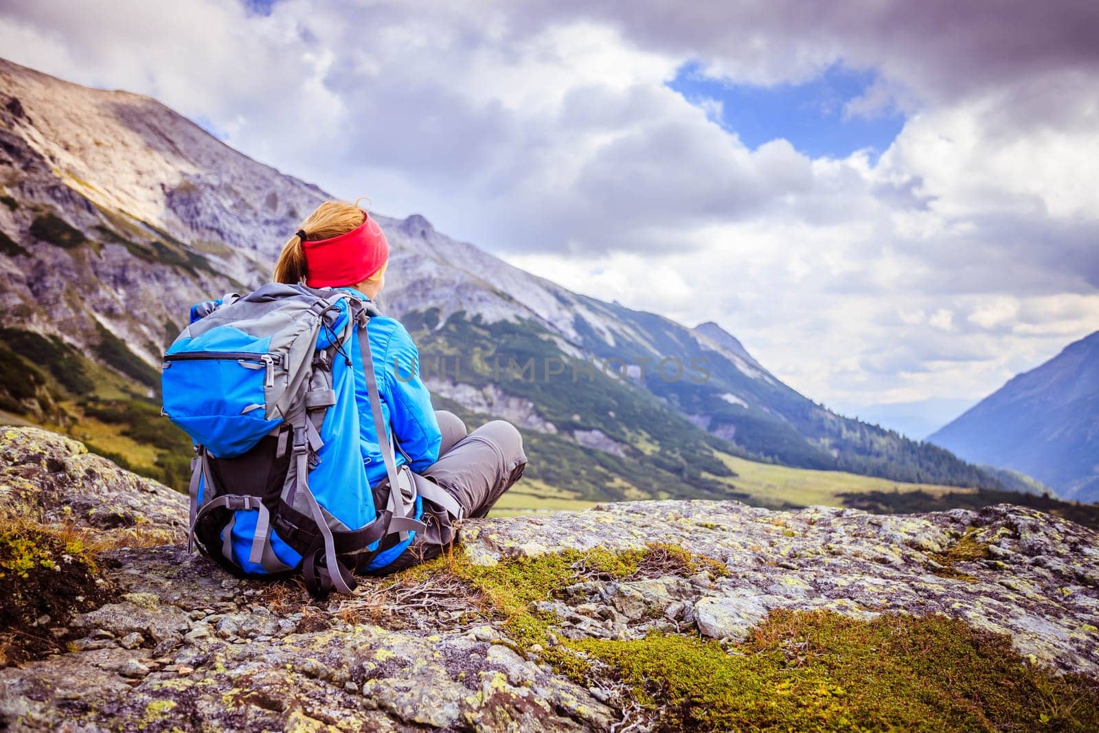 Woman in sportswear and with backpack is sitting on the stony ground and enjoys the view, in the mountains