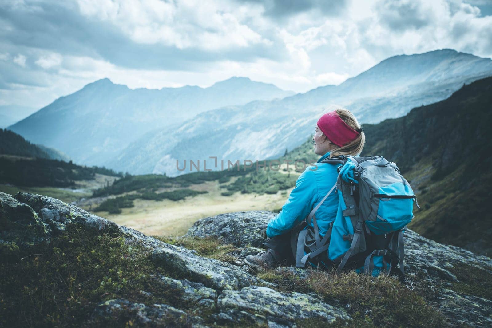 Woman in sportswear and with backpack is sitting on the stony ground and enjoys the view, in the mountains