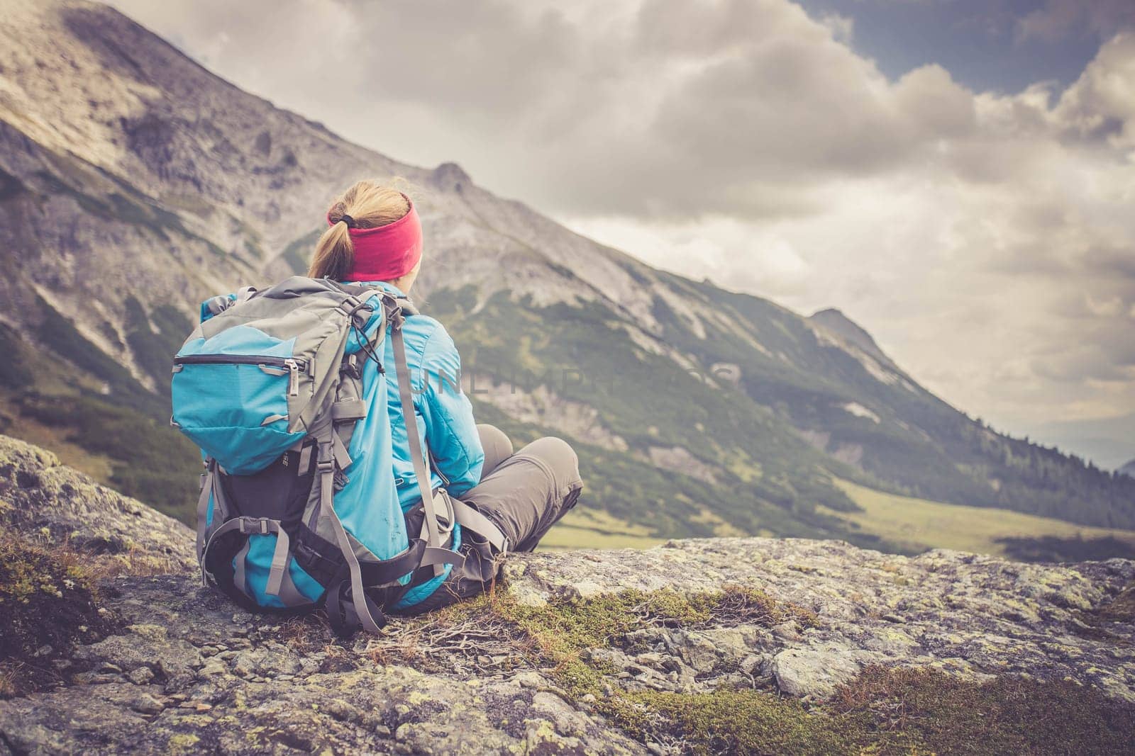 Woman in sportswear and with backpack is sitting on the stony ground and enjoys the view, in the mountains