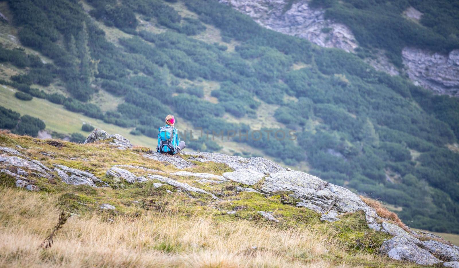 Woman in sportswear and with backpack is sitting on the stony ground and enjoys the view, in the mountains