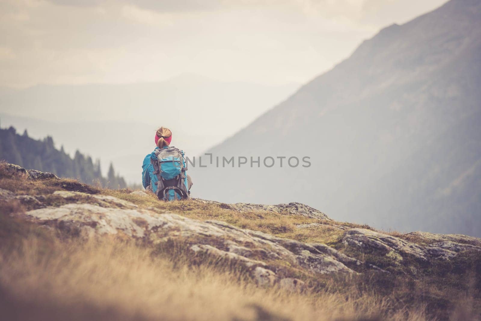 Woman in sportswear and with backpack is sitting on the stony ground and enjoys the view, in the mountains