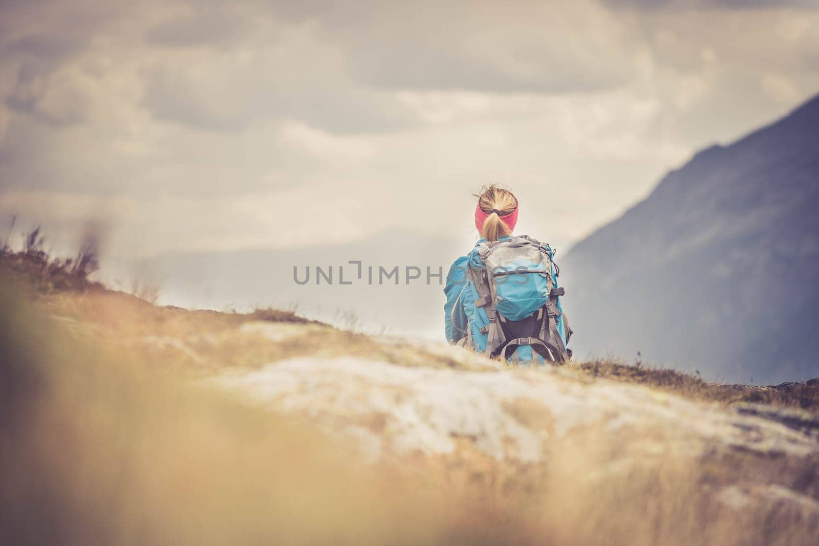 Woman in sportswear and with backpack is sitting on the stony ground and enjoys the view, in the mountains