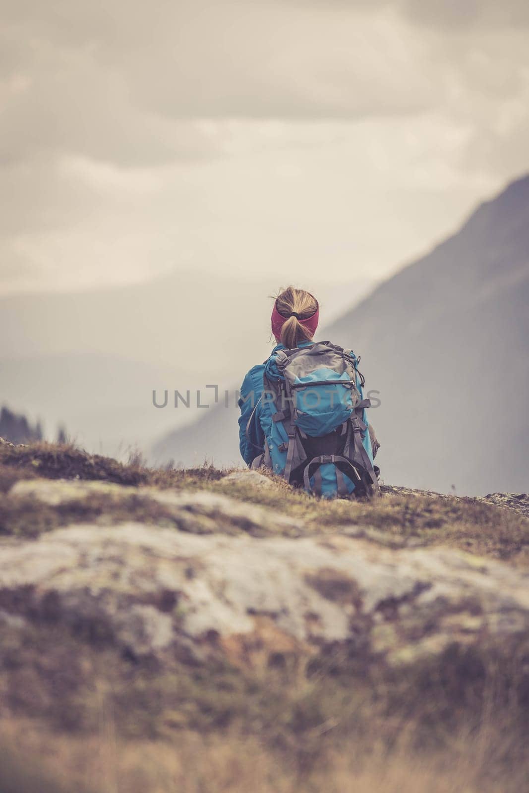 Woman in sportswear and with backpack is sitting on the stony ground and enjoys the view, in the mountains