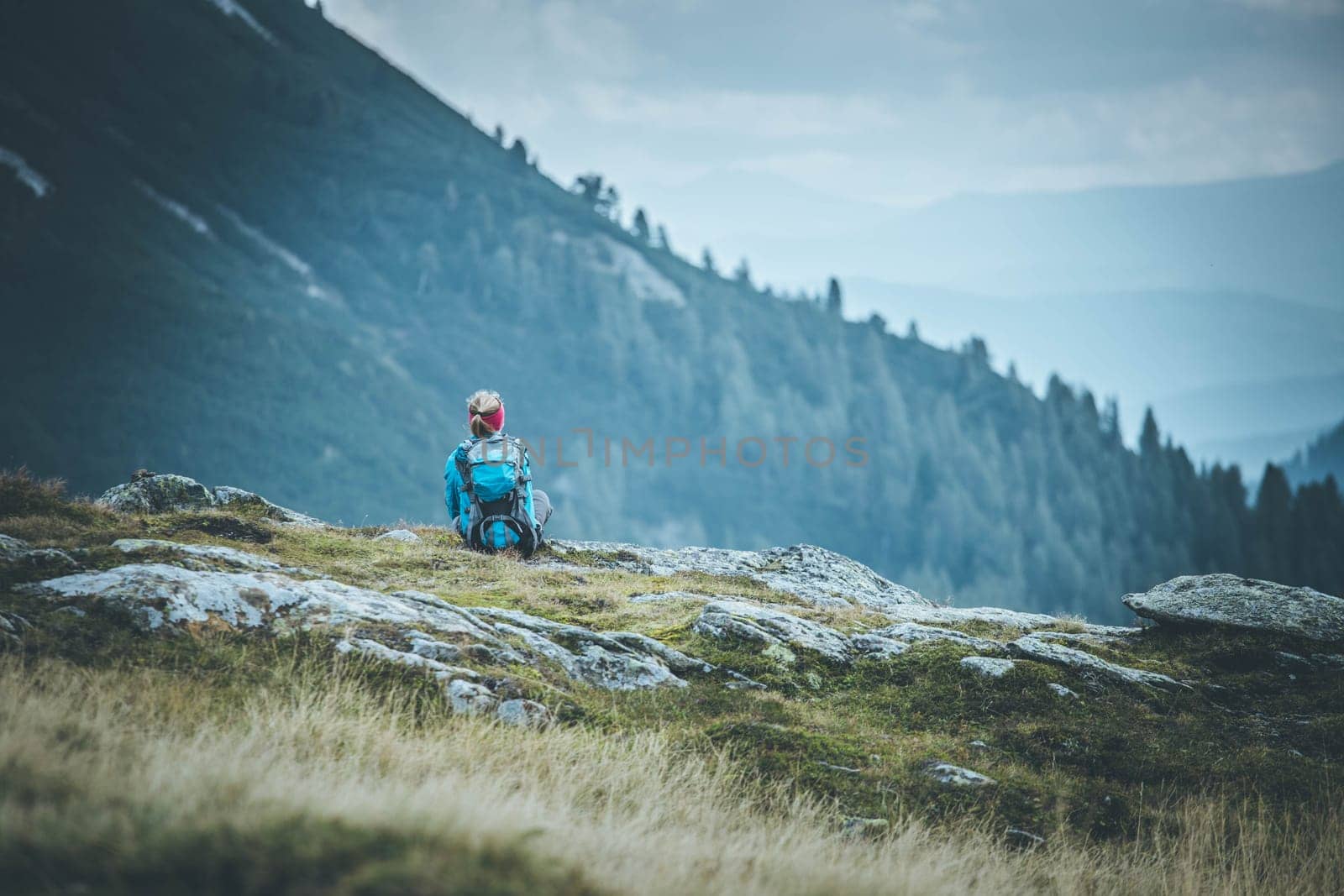 Woman in sportswear and with backpack is sitting on the stony ground and enjoys the view, in the mountains