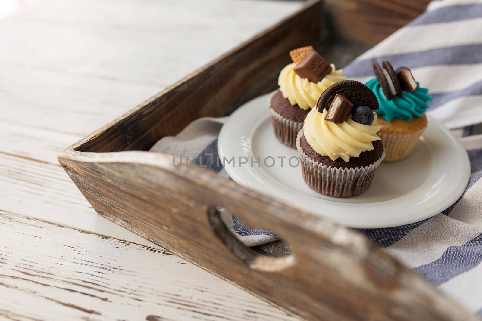 Tasty multicolored muffins and cupcakes on the white plate on a wooden tray with towel. Decorated with different candies, cookies and colored sweet cream cheese on top. High quality photo