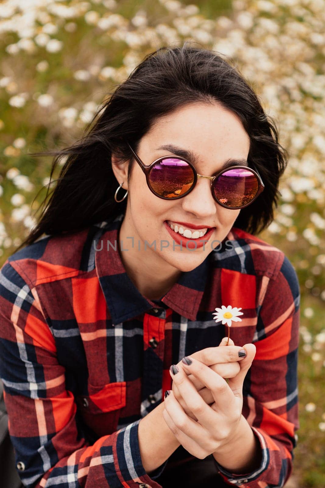 Beautiful woman with sunglasses sitting in the middle of a flowery meadow smelling a daisy