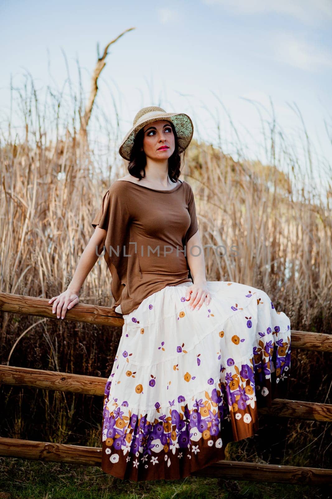 Stylish woman with long skirt sitting on the top of a wooden fence in the field