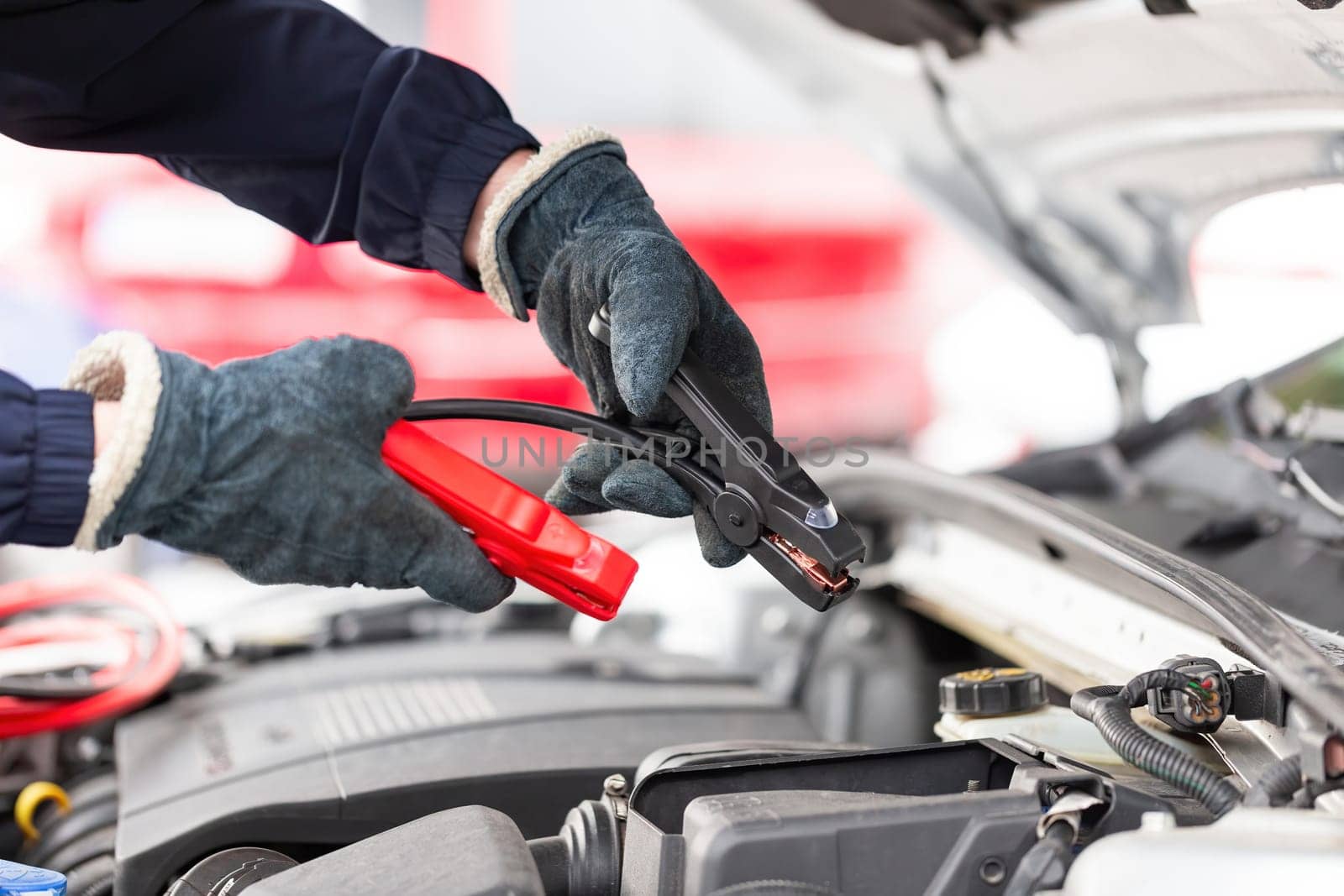 Human trying to start engine with jumper cables in close up. Emergency vehicle start-up. Hands in winter gloves holding black and red tool over open hood of a car.