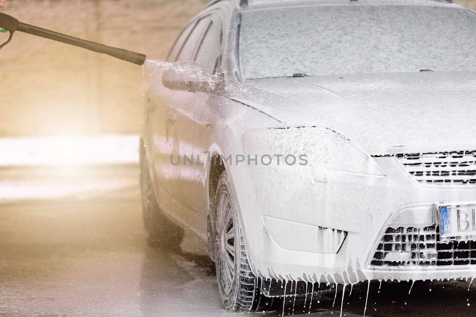 High pressure automobile cleaning with foam in carwash. Washing car by using soap and water nozzle. Vehicle purifying in self-service washbasin on sunny day.
