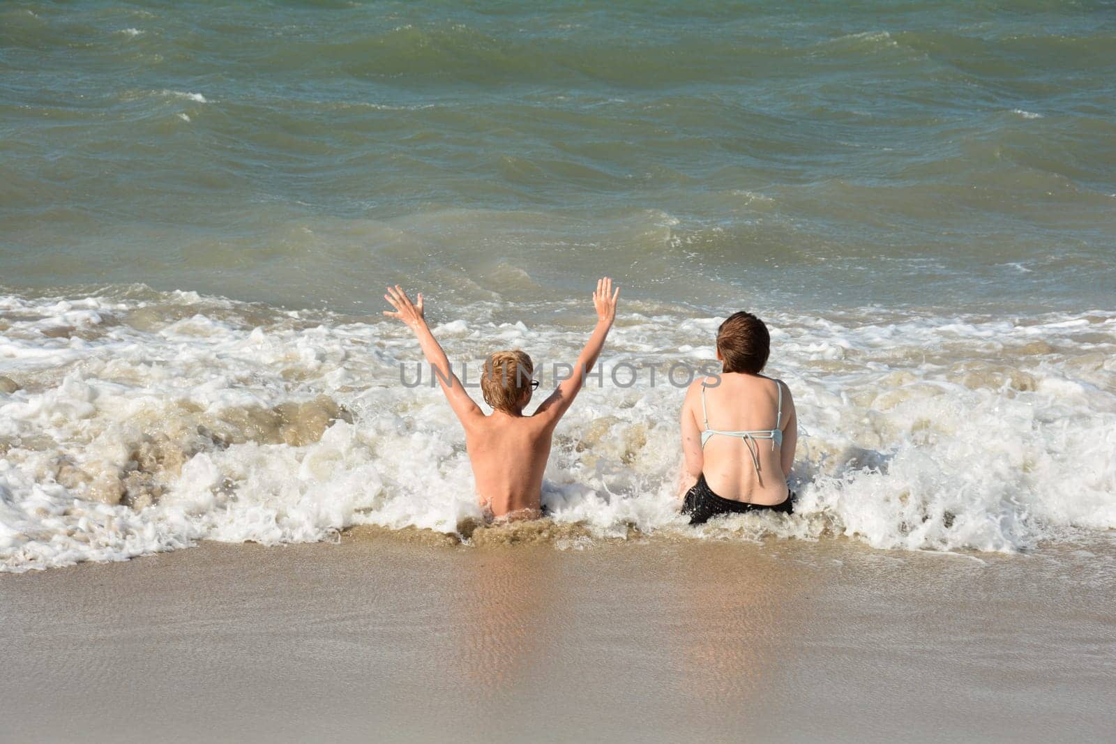 Two children sit full of joy on the sandy beach in the middle of the waves by the sea, on the North Sea coast in the Netherlands, Zeeland