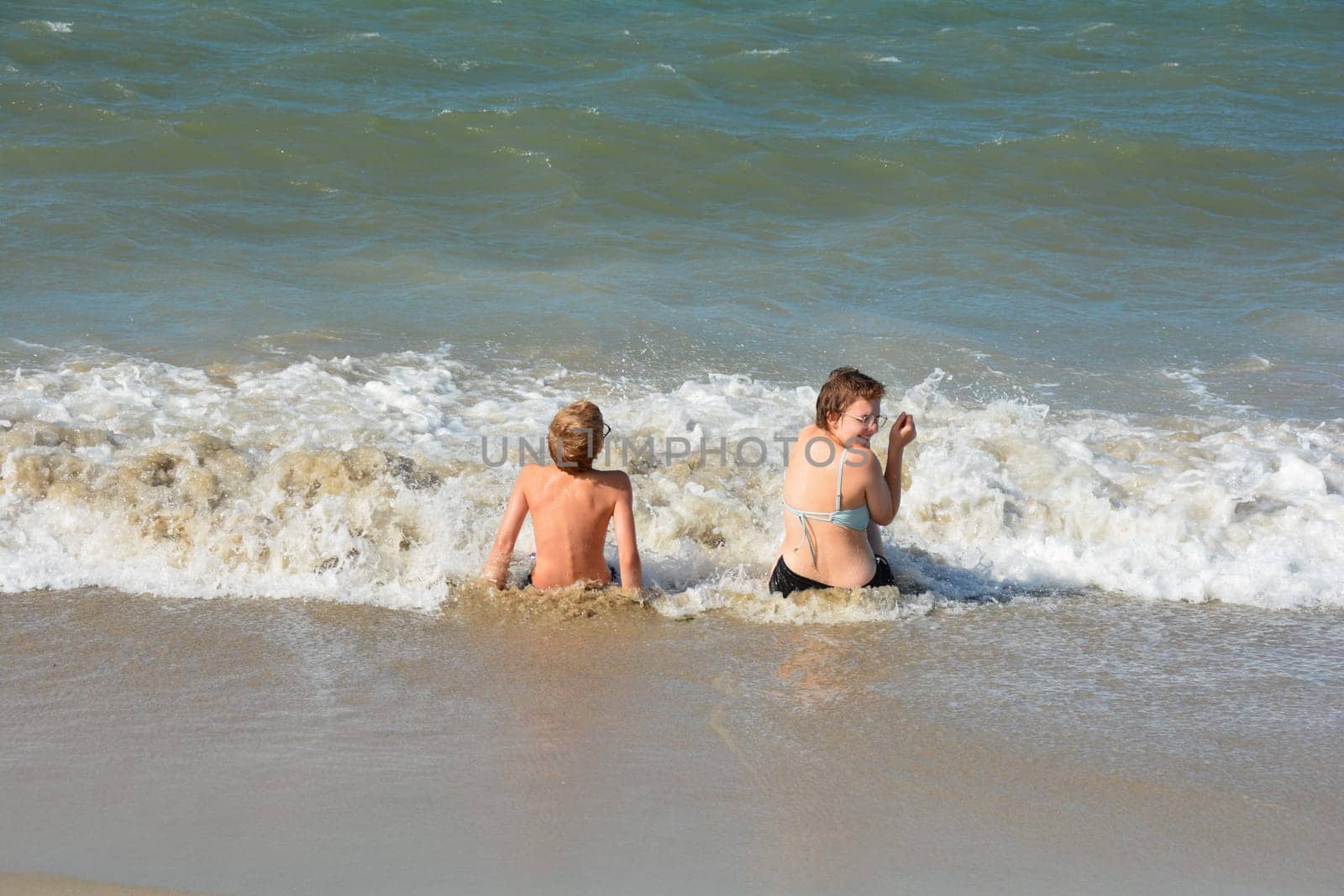 Two children sit full of joy on the sandy beach in the middle of the waves by the sea, on the North Sea coast in the Netherlands, Zeeland