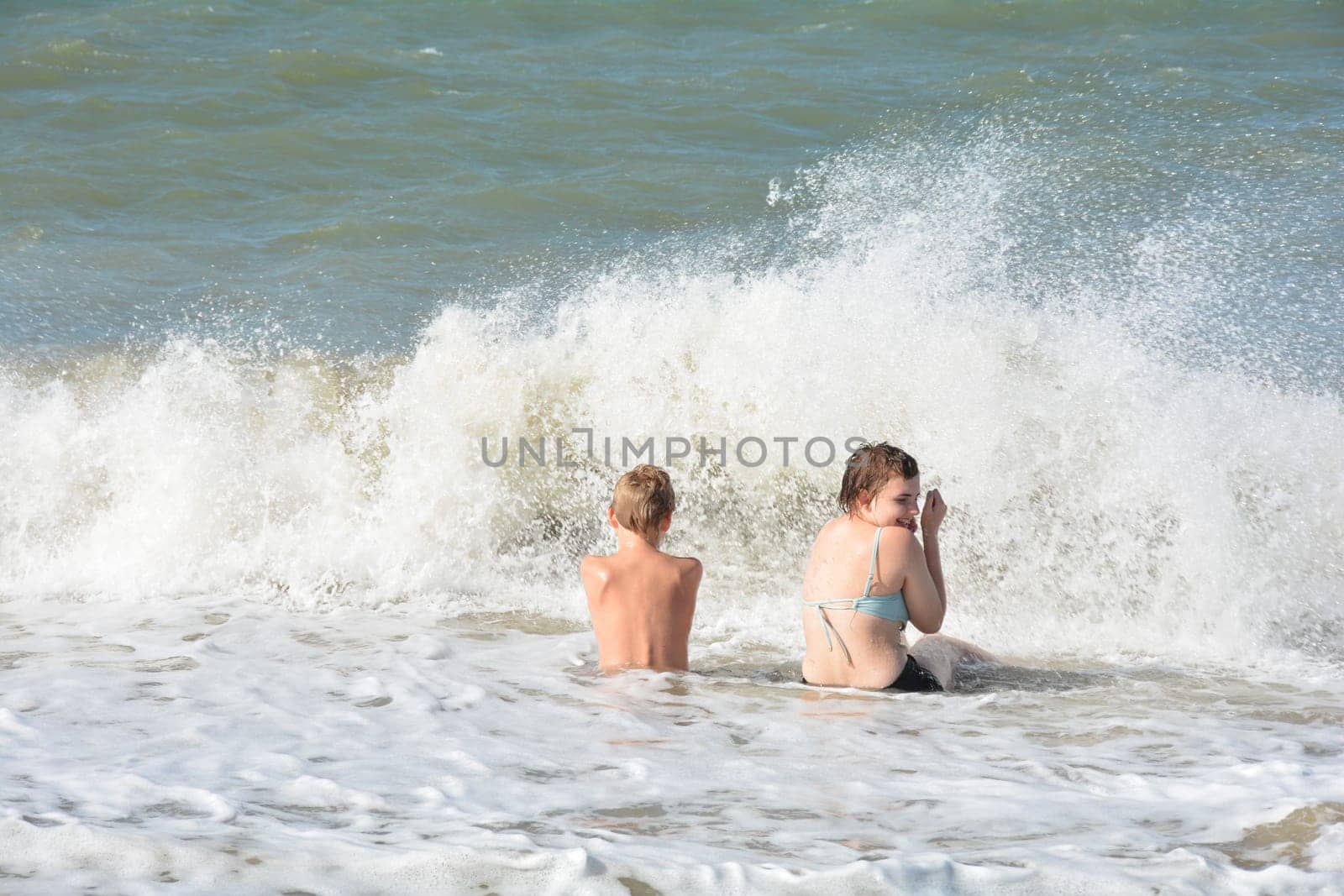 Two children sit full of joy on the sandy beach in the middle of the waves by the sea, on the North Sea coast in the Netherlands, Zeeland