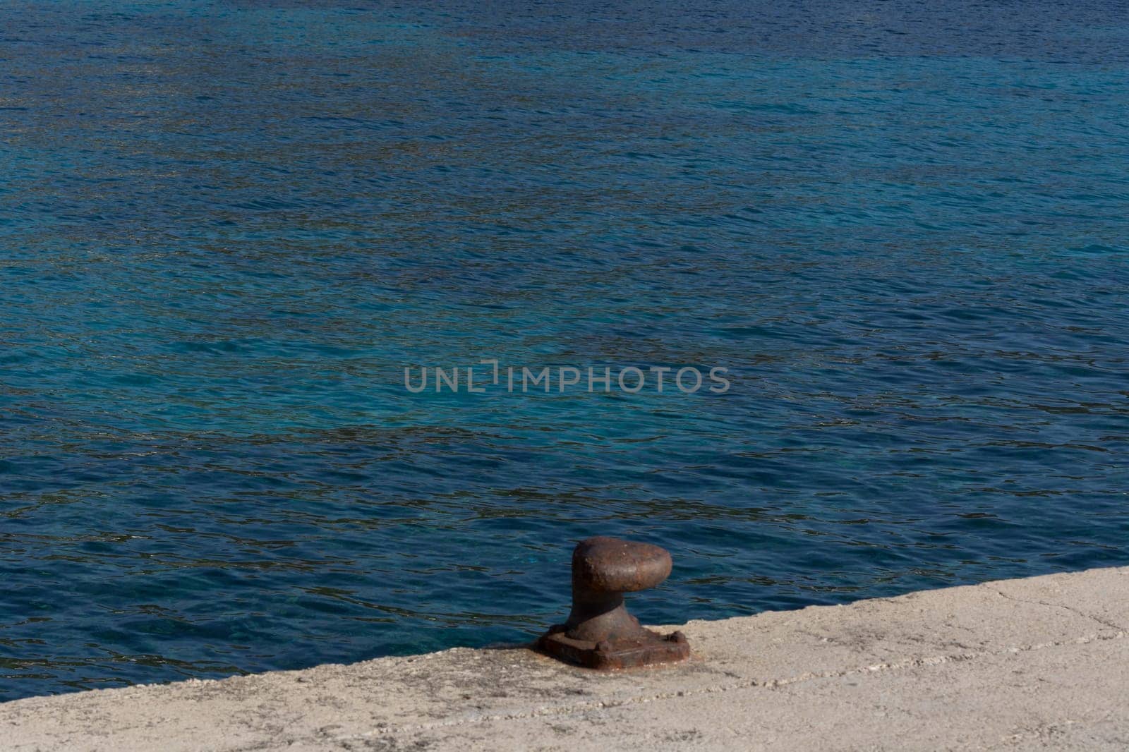 A rusty bollard on a quay wall with a rusty chain and in the background the water in the harbor on the Mediterranean Sea.