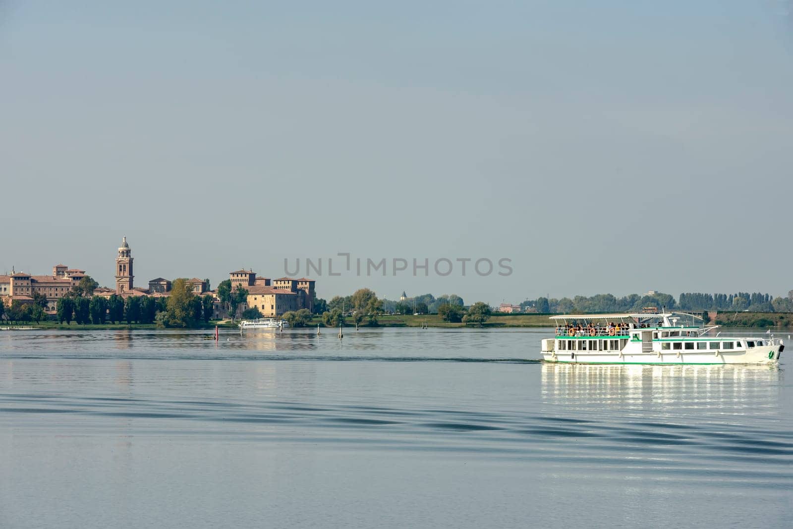 view of touristic passenger vessel cruising on Mincio lake with Gonzaga Ducale Palace in background, shot in bright autumn sun light  at Mantua, Lombardy, Italy.