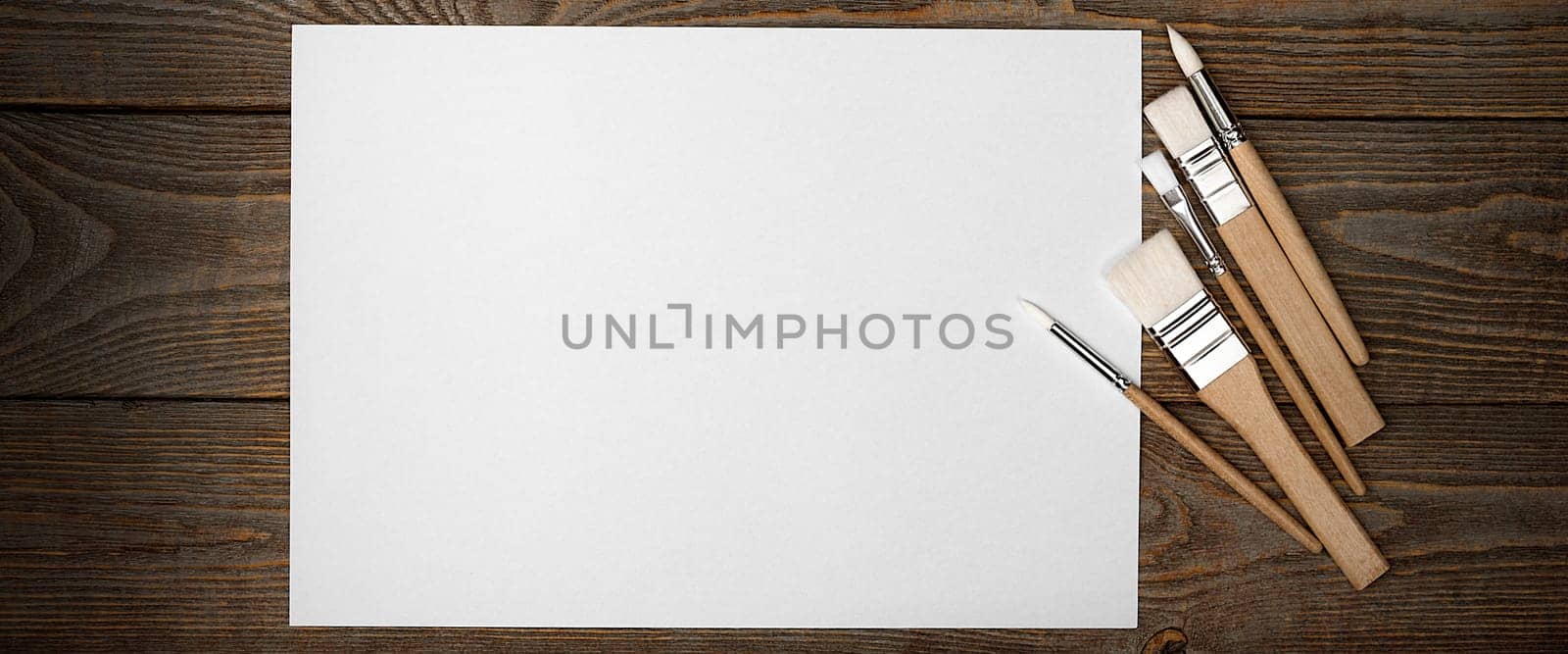 A clean white sheet and brushes on a wood textured background with space to copy. Layout, mockup space for text and drawing.