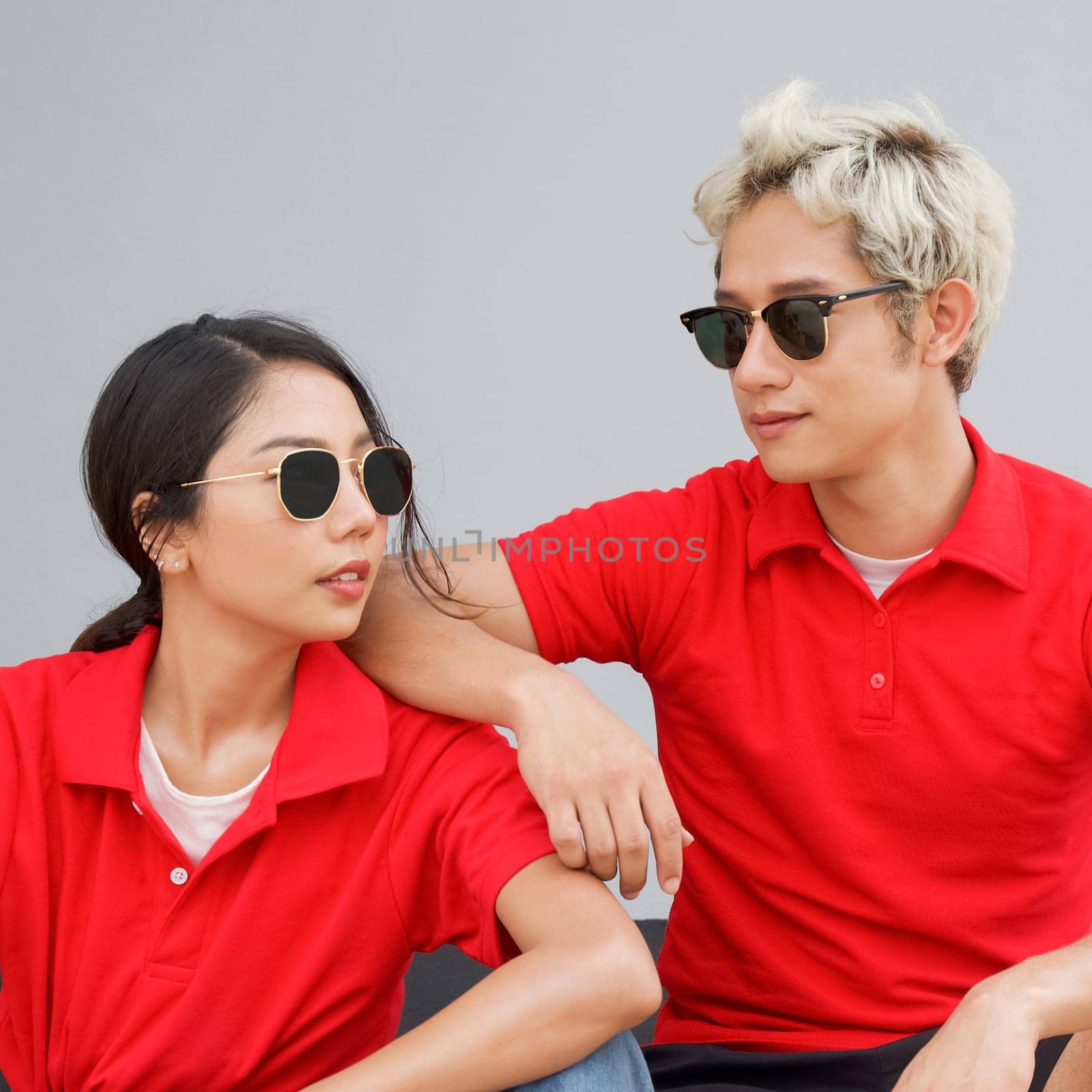 Young asian couple in red T-shirt with collar and black sunglasses looking at each other. Portrait on gray background.