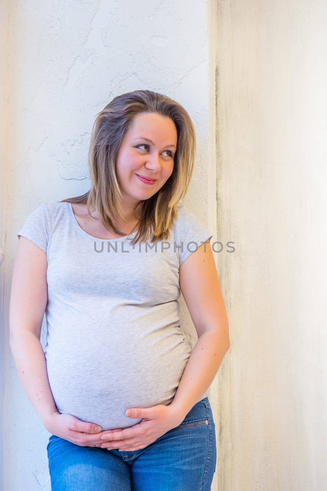 Beautiful pregnant woman standing near window at home in light interior and holdinh her tummy