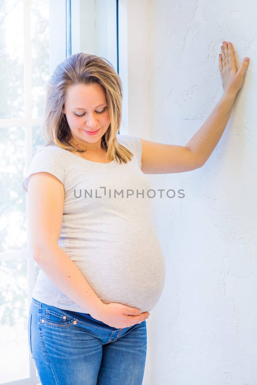 Beautiful pregnant woman standing near window at home in light interior and holdinh her tummy