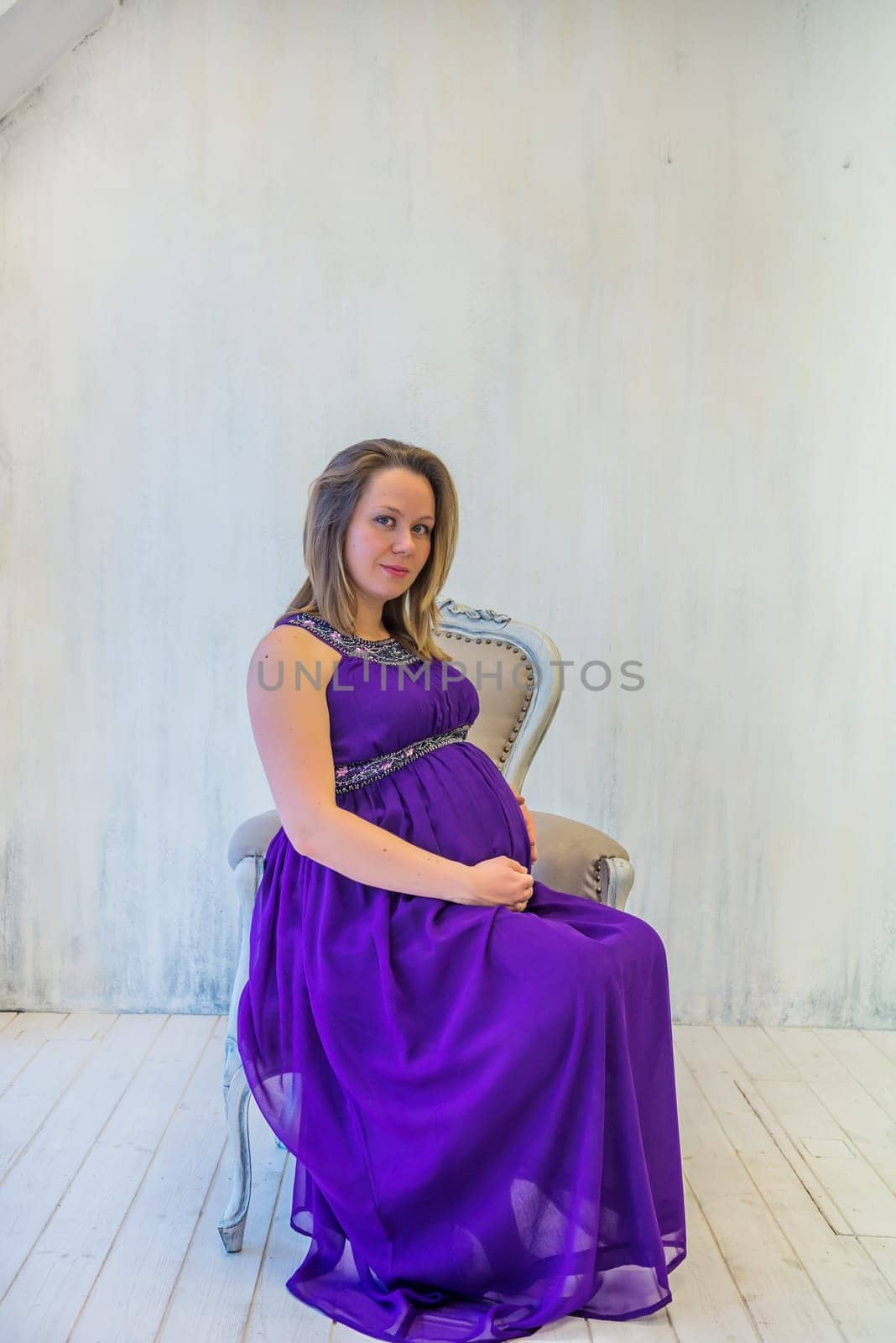 Beautiful pregnant woman in ultra violet dress sitting on the armchair in light interior