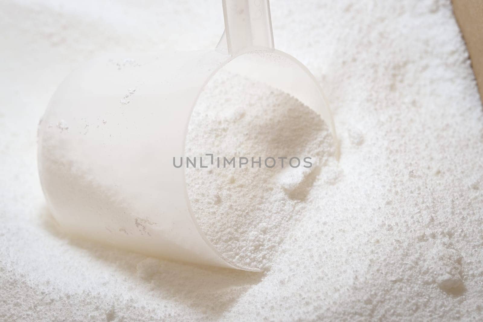 Close up of clear plastic scoop being used to measure white laundry detergent powder in a cardboard box. Bright image with macro powder detail