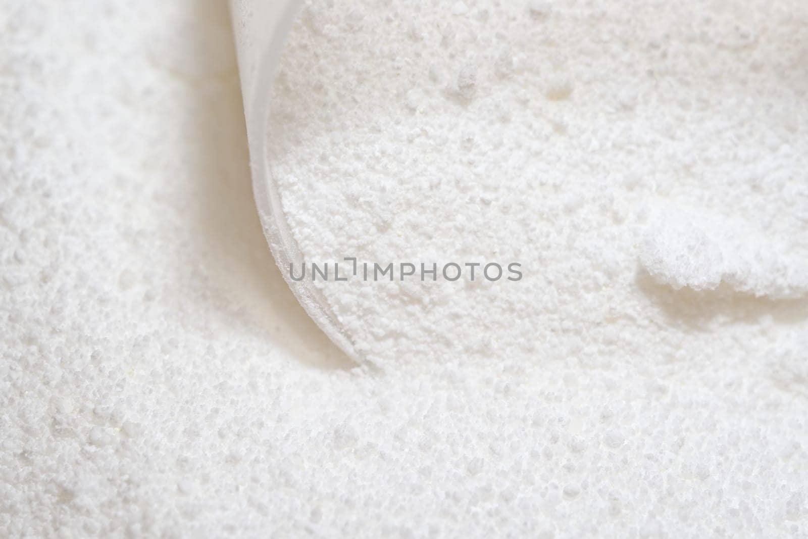 Pure white laundry detergent powder, clumped together and being picked up by a clear plastic measuring scoop. Bright image and super macro perspective