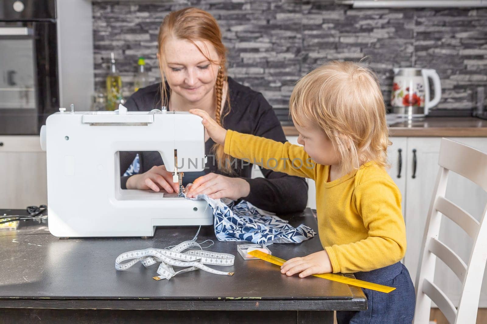 Smiling young mother is sewing and her son is holding sewing machine. Horizontally.
