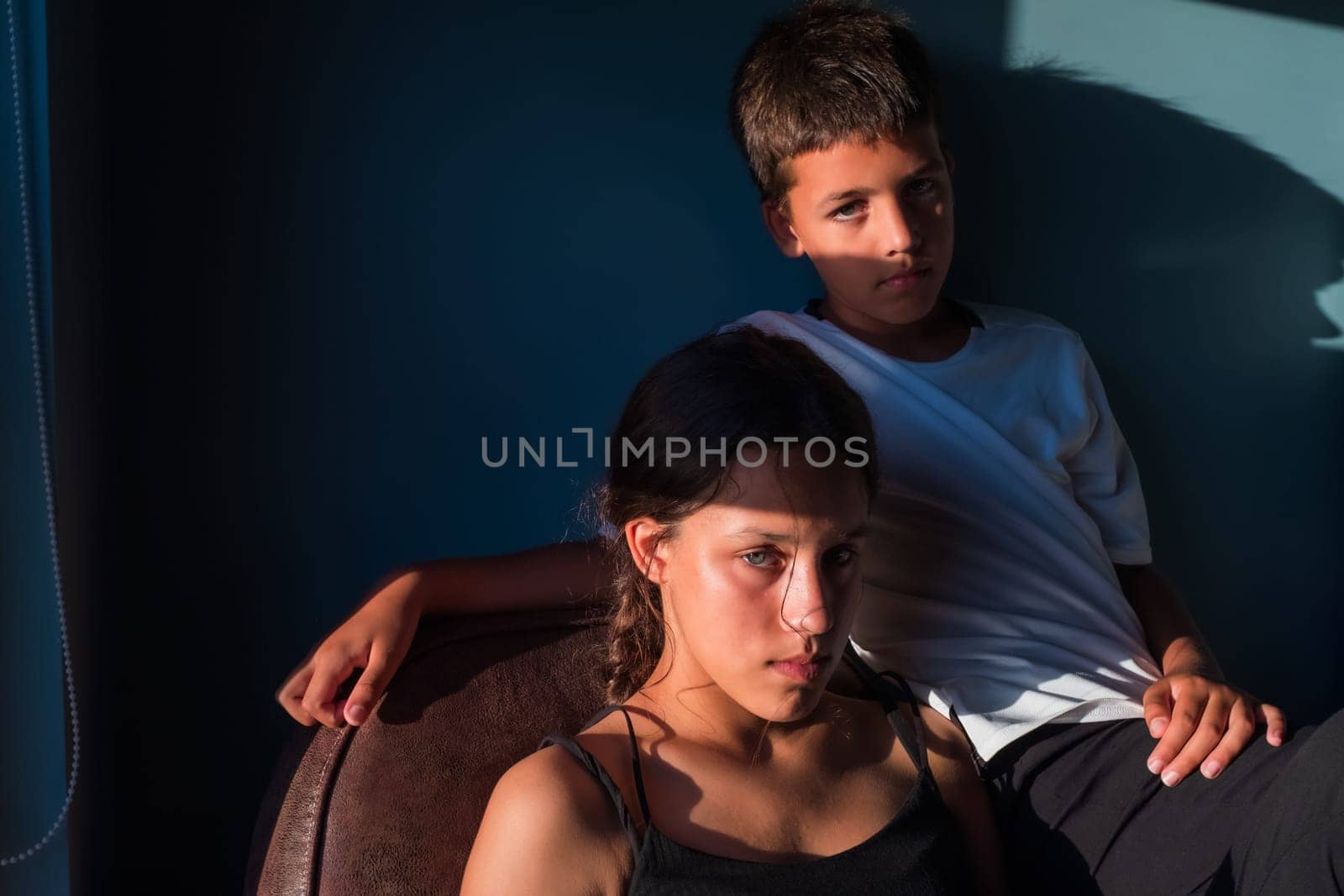 Portrait of brother and sister sitting inside looking at camera with serious expression. Source of natural light that highlights the same color of the two brothers' eyes.