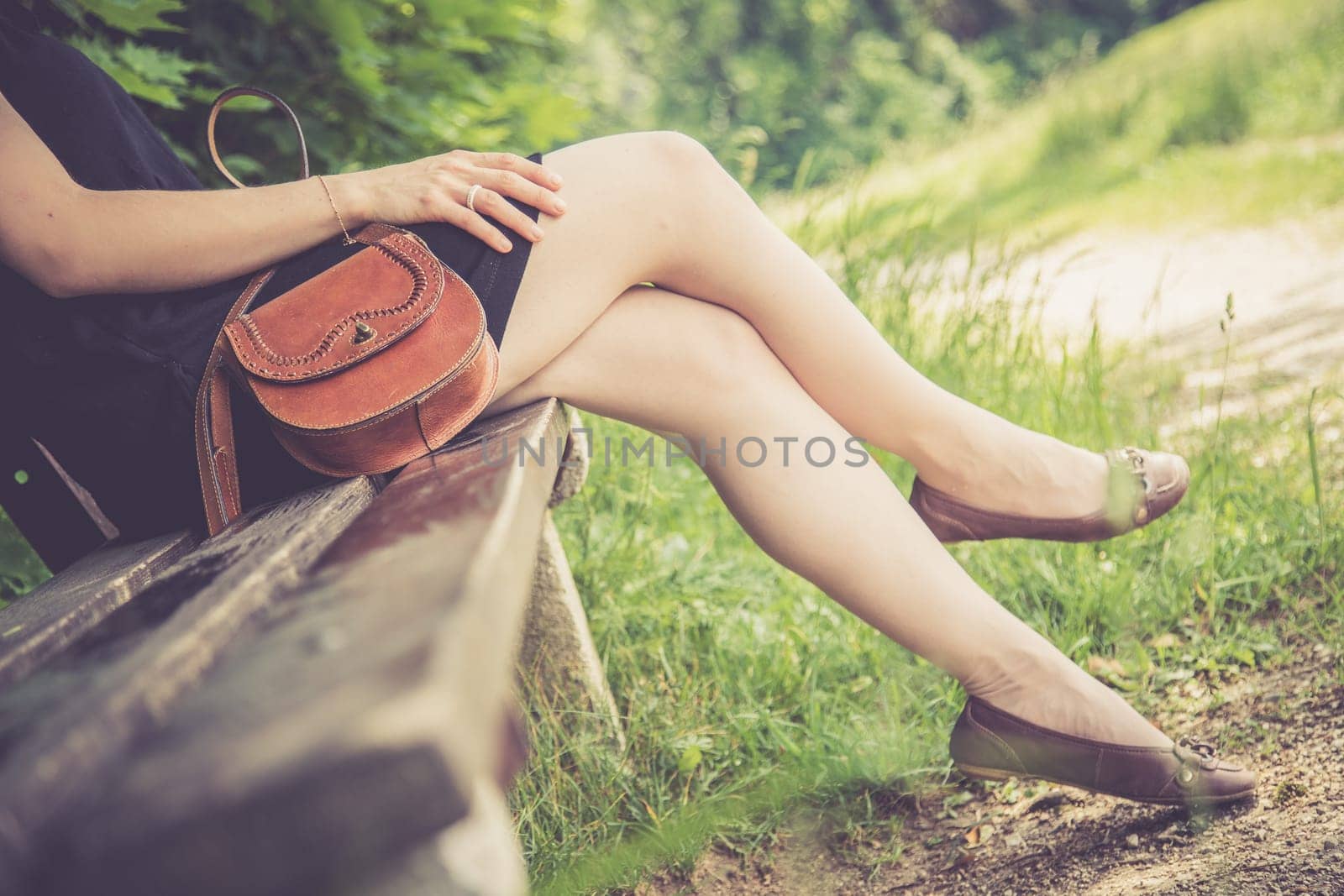 Young woman in black dress with sexy legs and leather handbag is sitting on a park bench, summer