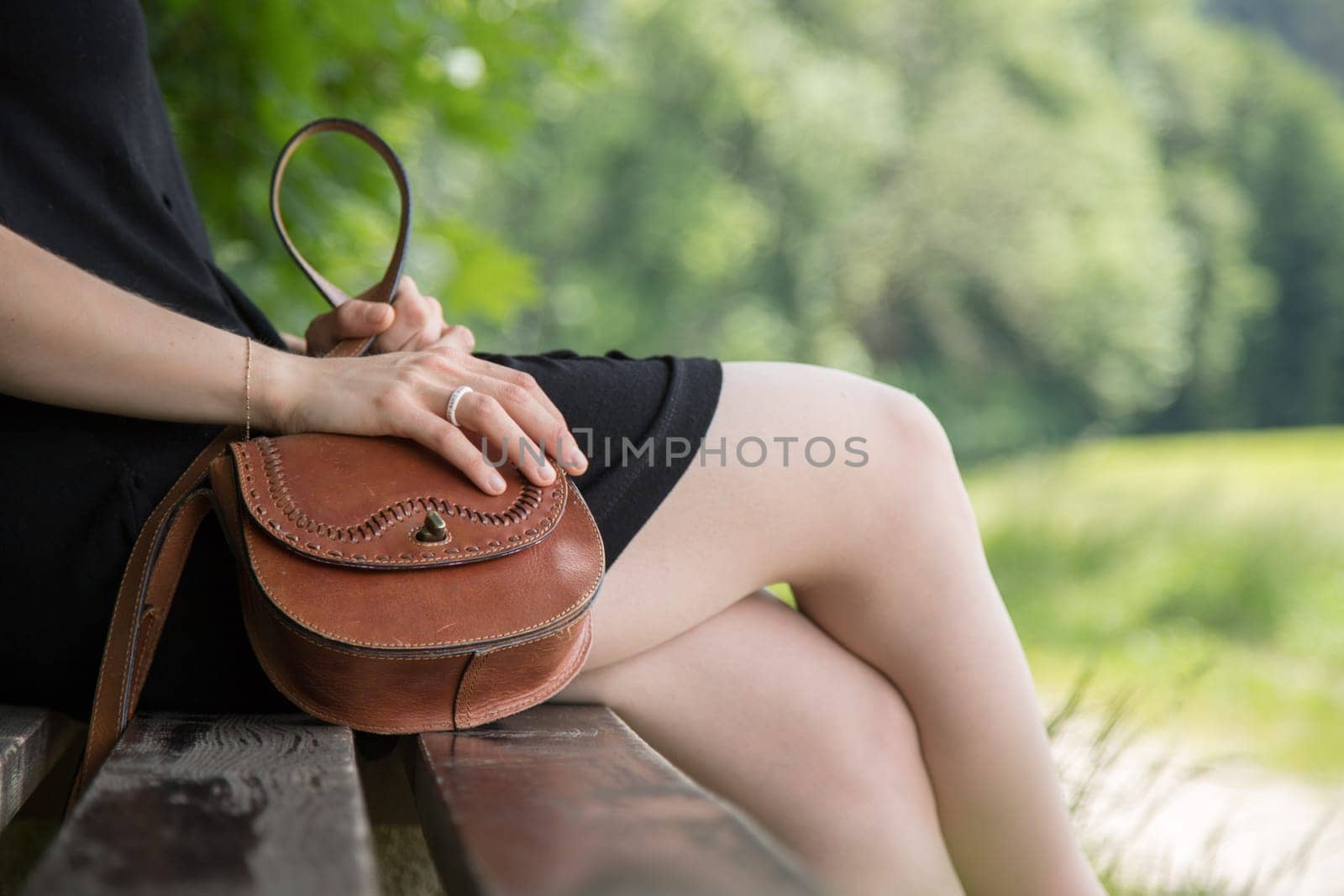 Young woman in black dress with sexy legs and leather handbag is sitting on a park bench, summer