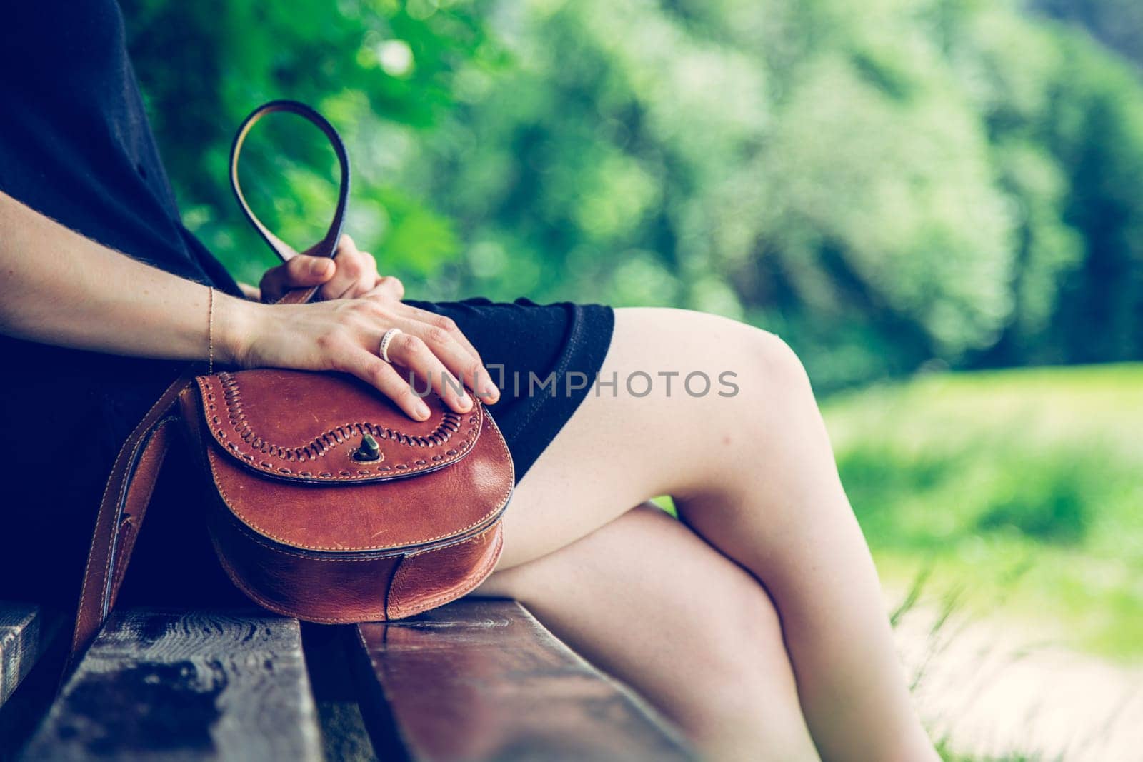 Young woman in black dress with sexy legs and leather handbag is sitting on a park bench, summer