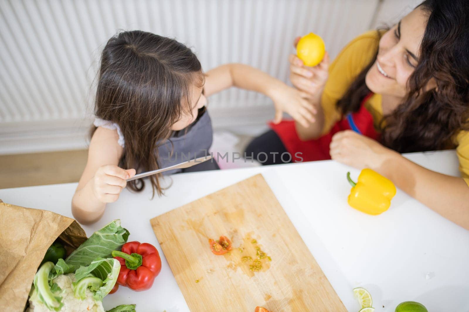 Happy little girl and her mother slicing lemons and tomatoes on cutting board. Smiling woman and young child at white talbe holding lemons. Daughter-mother cooking together