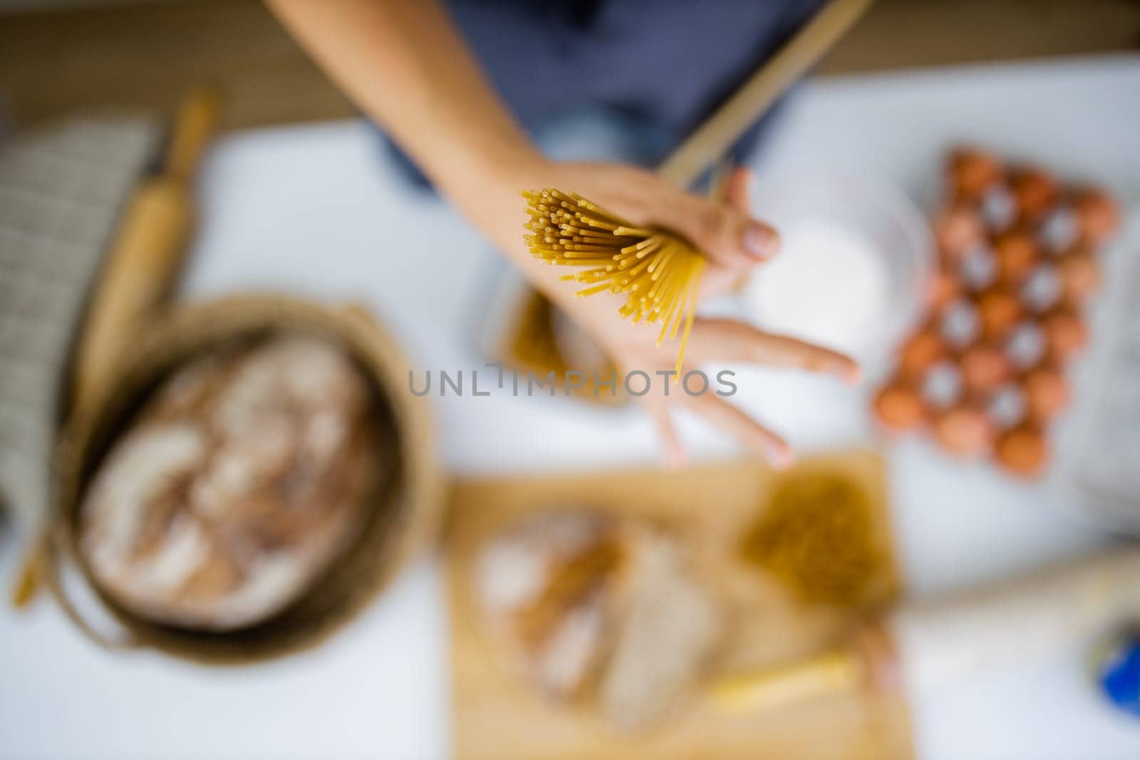 Female hand holding uncooked spaghetti above blurry ingredients on white table. Blurry eggs, pasta, flour, and bread on table from above. Pasta preparation