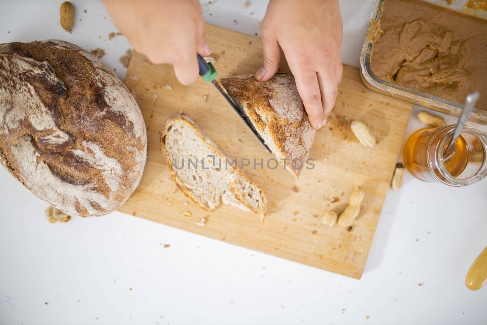 Female hands slicing bread on wooden cutting board from above. Woman cutting bread next to peanut butter and a honey jar. Healthy meal preparation