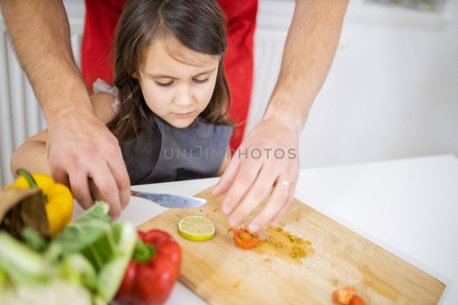 Little girl and her father slicing lemons, tomatoes, and bell peppers on cutting board. Male hands and young child cutting vegetables on wooden board. Daughter-father cooking together