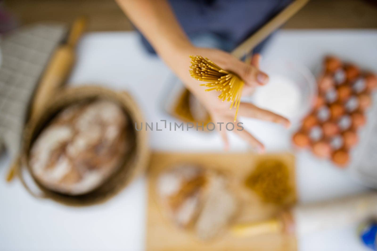 Female hand holding uncooked spaghetti above blurry ingredients on white table. Blurry eggs, pasta, flour, and bread on table from above. Pasta preparation