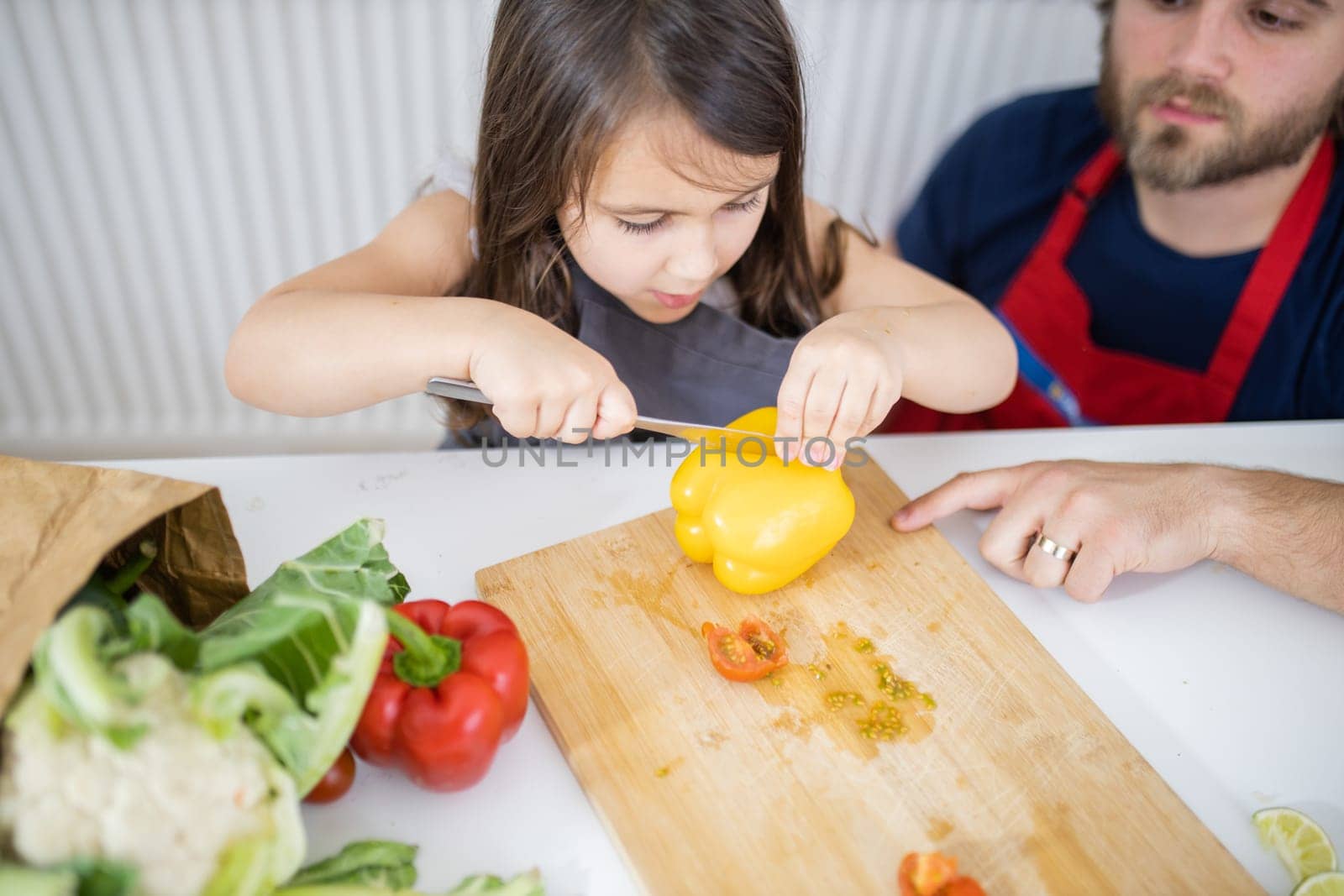 Little girl and her father slicing yellow bell pepper on cutting board. Man helping young child cutting vegetables on wooden board. Daughter-father cooking together