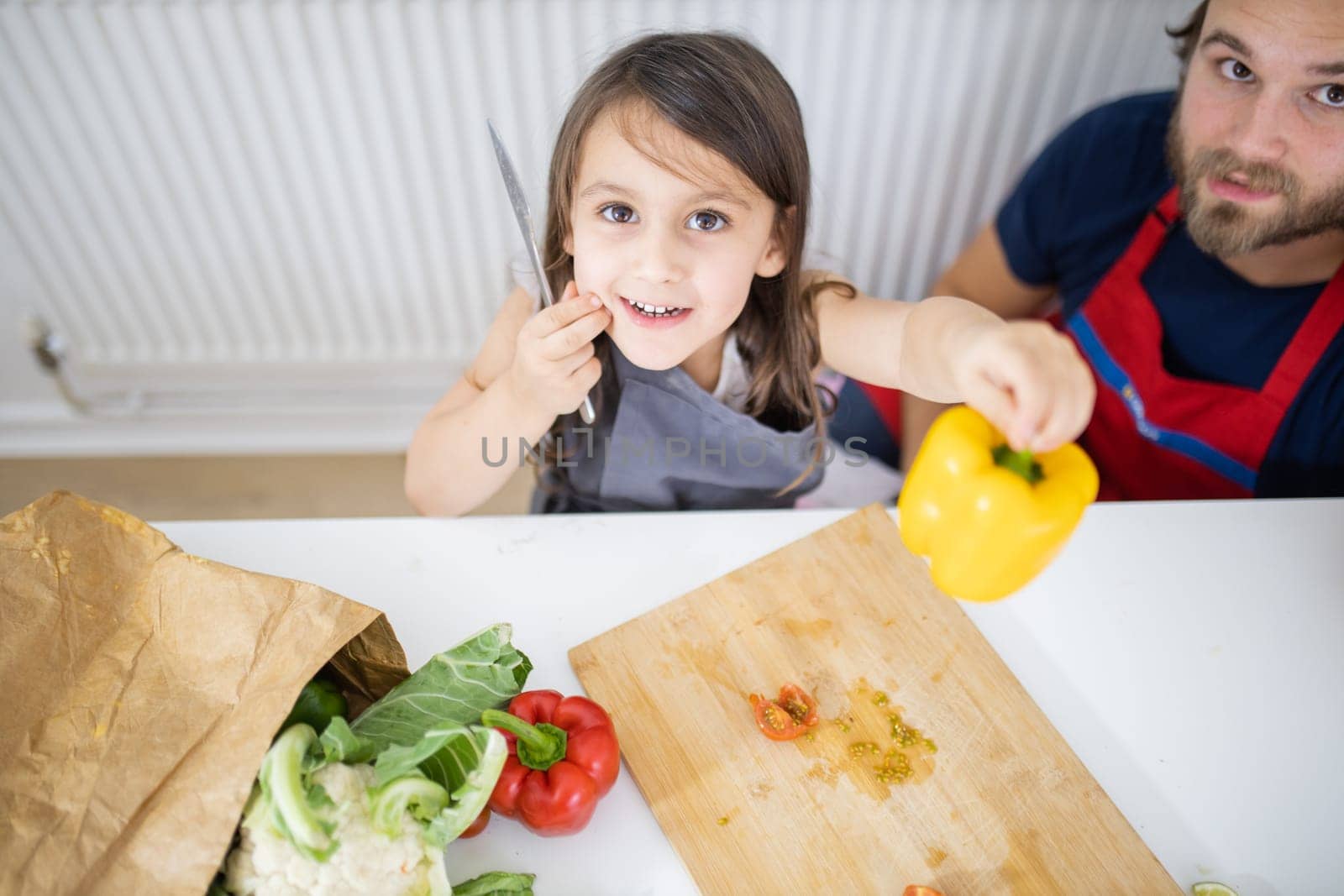 Happy little girl and her father showing yellow bell pepper above cutting board. Man and young child cutting vegetables on wooden board. Daughter-father cooking together