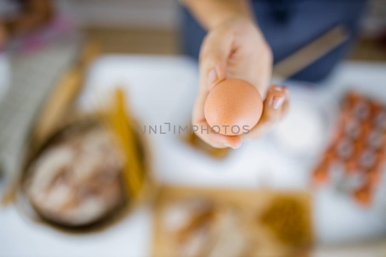 Female hand holding uncooked egg above blurry ingredients on white table. Blurry pasta, eggs, flour, and bread on table from above. Healthy meal preparation